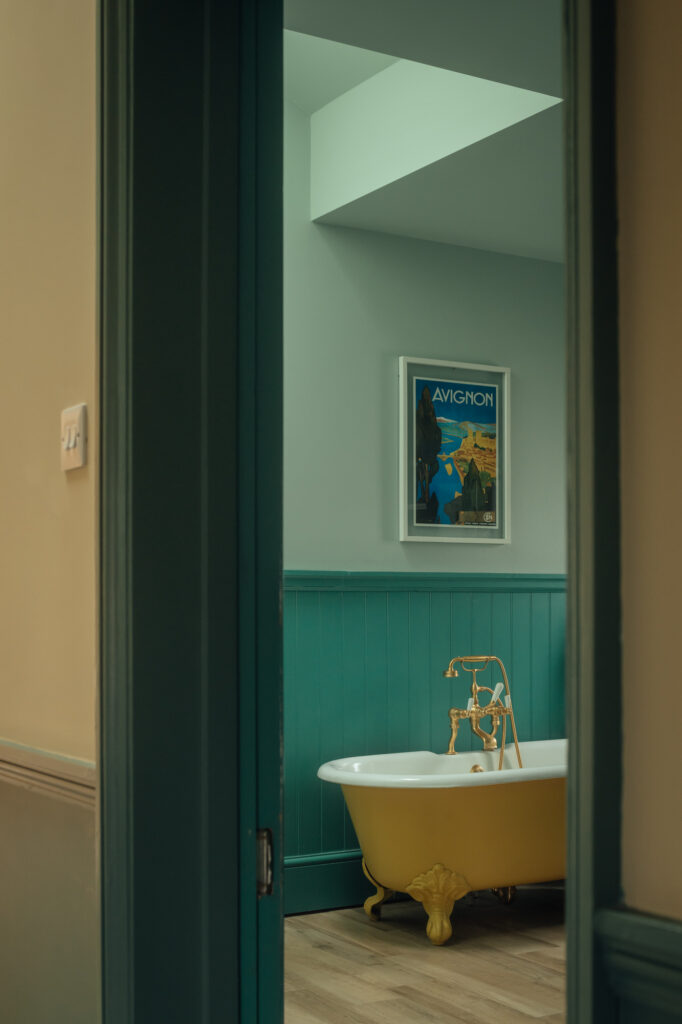 View into a bathroom with a yellow clawfoot bath and gold fittings against teal wainscotting and timber cladding, finished with a framed travel poster of Avignon on the wall.