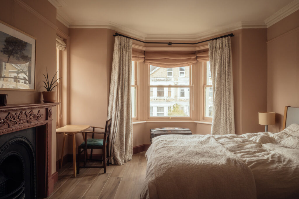 A bedroom with a bed, a small wooden desk and chair, and a fireplace, featuring a bay window with timber cladding looking out onto a residential street.