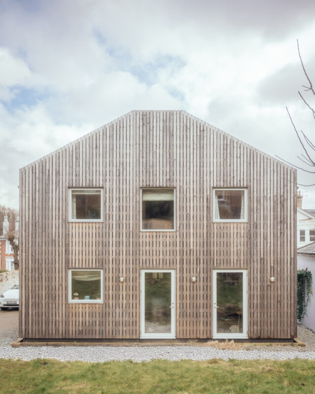 A two-storey house with a gabled roof and vertical timber cladding, featuring symmetrical windows and two glass doors on the ground floor.