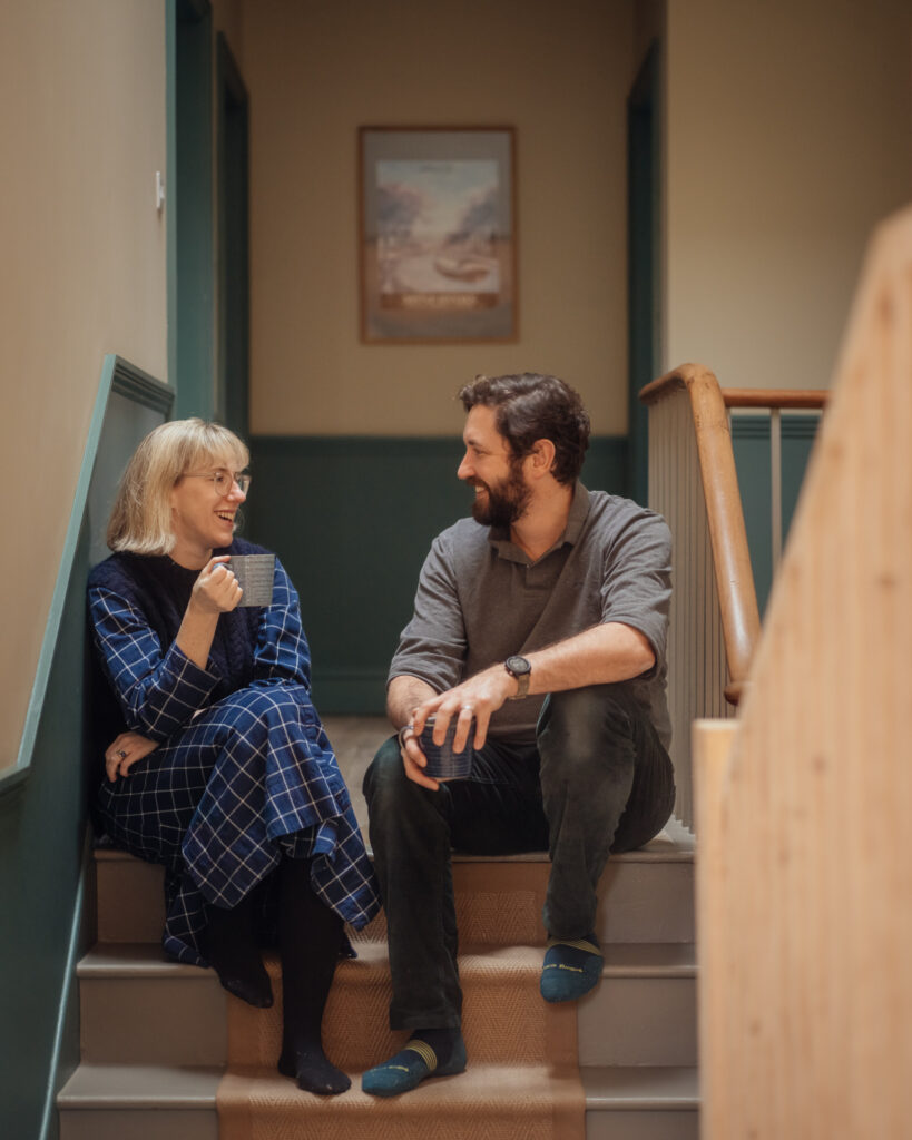 A woman and a man sit on a staircase in a timber structure, laughing and holding mugs of coffee.