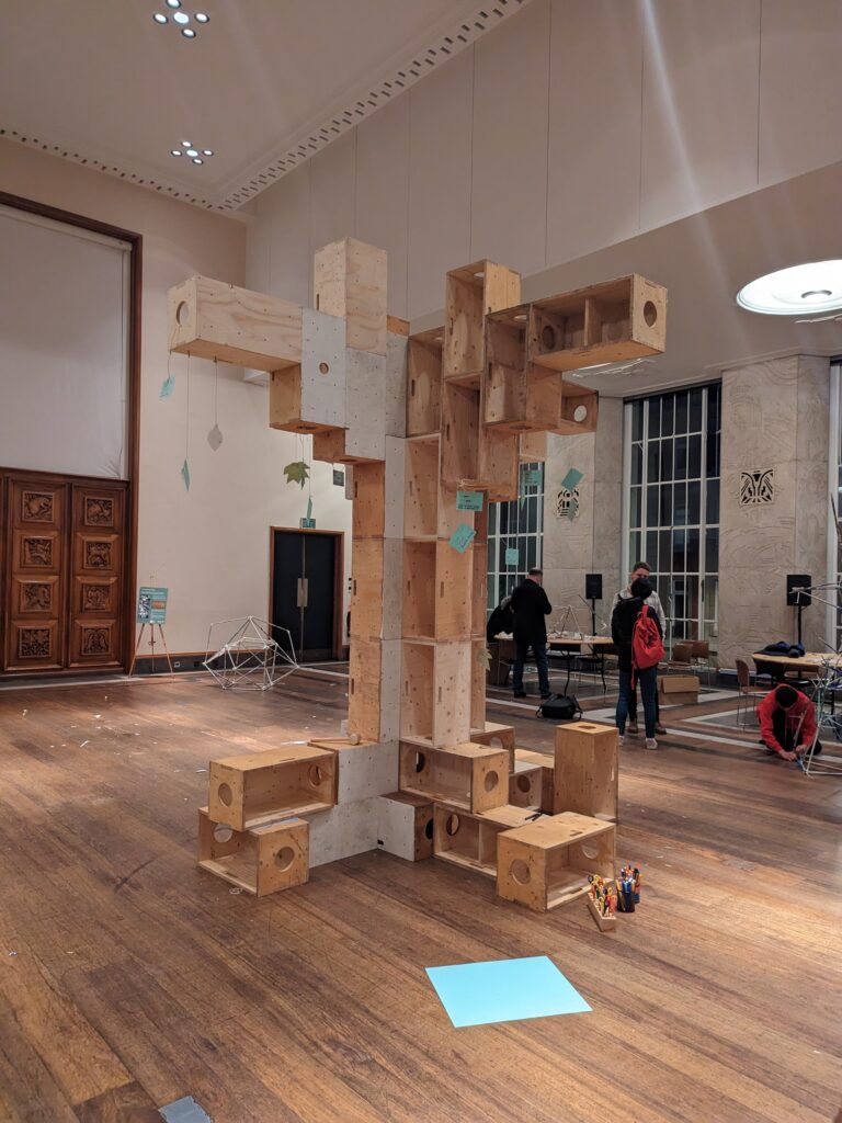 A large abstract tree sculpture made of reused plywood boxes stands in a spacious gallery. Created as part of a community build project, this striking structure embodies youth architecture and design. People observe in the background, whilst art supplies for the make-and-build process lie on the floor near the sculpture.