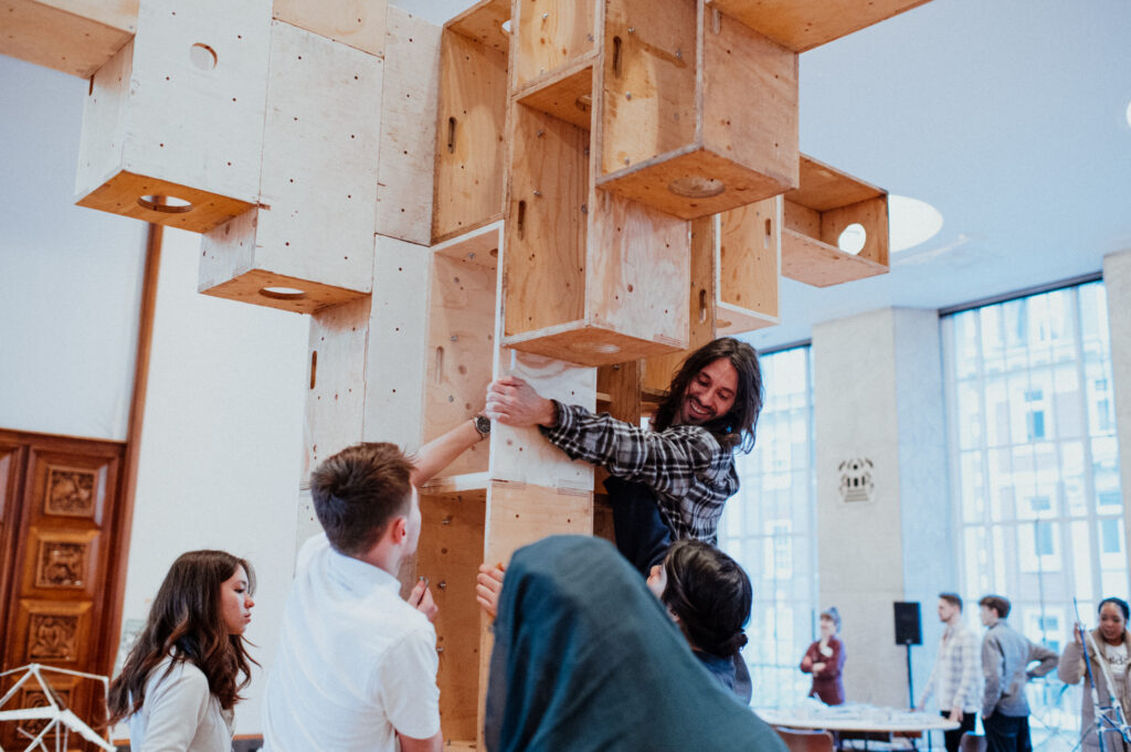 A group of people participate in a Community Build Project, assembling large plywood cubes into a tall structure indoors, working together and smiling. Bright windows and tables are visible in the background, highlighting a creative atmosphere where youth architecture design and the spirit to make and build are celebrated.