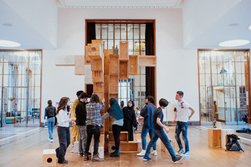 A group of people interact with a large, modular birch structure in a spacious, modern room with high ceilings, wooden floors, and large windows letting in natural light.