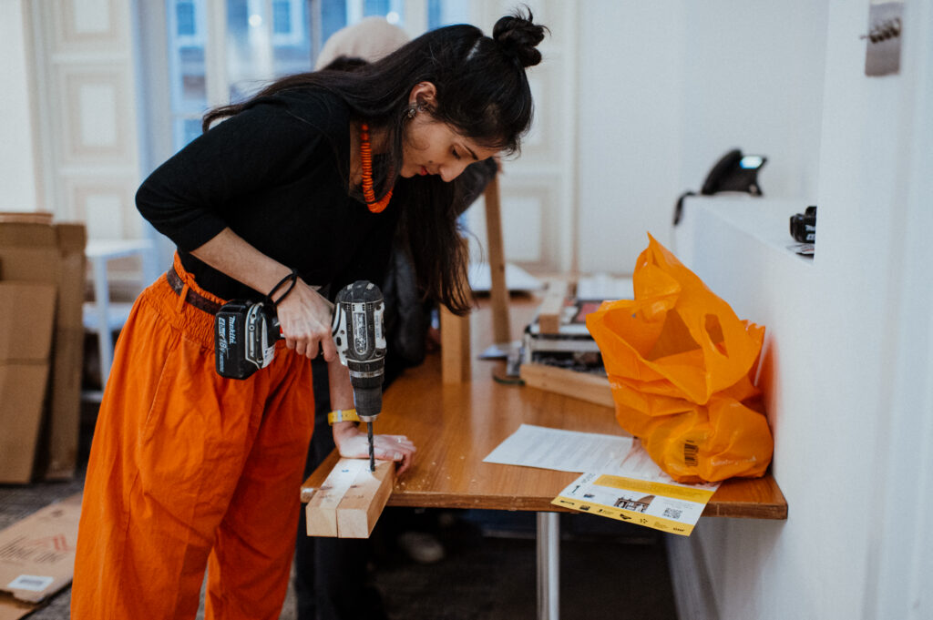 A woman in orange trousers uses a power drill on a wooden plank placed on a table, contributing to a youth architecture design community build project. Papers, an orange bag, and tools are scattered across the table in the well-lit room, suggesting an engaging hands-on environment focused on making and building creative structures.