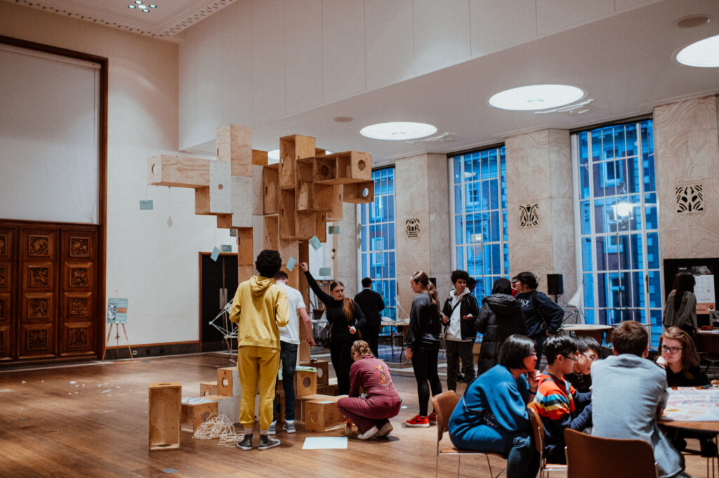People are gathered in a spacious room with large windows, assembling a tall plywood structure. Others sit at tables working on projects, suggesting a collaborative Community Build Project focused on youth architecture and design, where participants make and build innovative sculptures from reused materials.