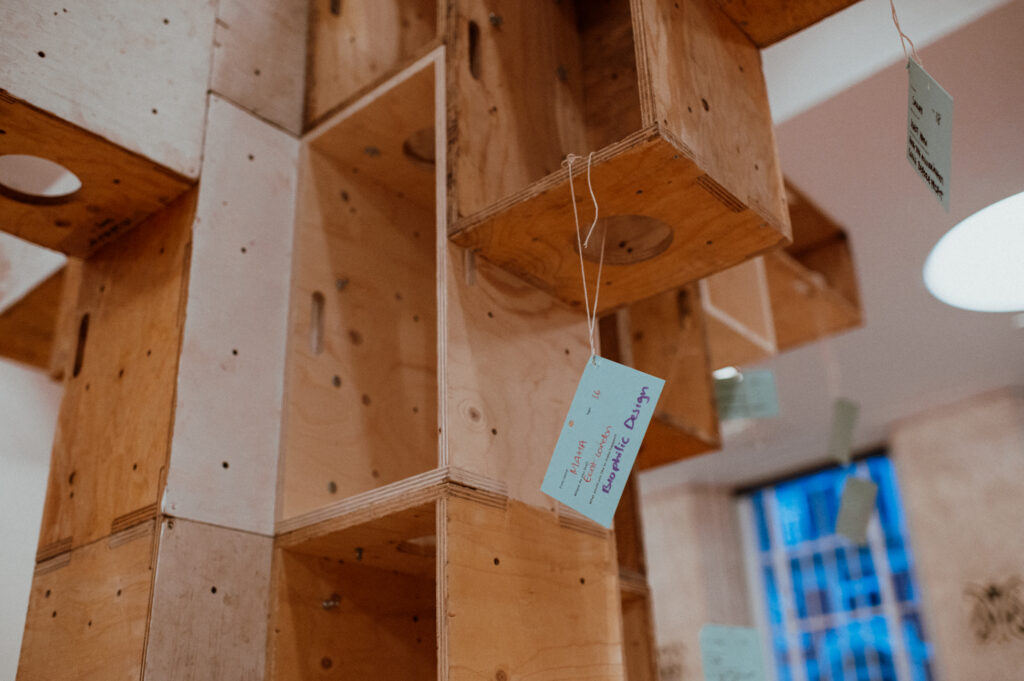 Close-up of a wooden structure crafted from plywood, with open cubbyholes featuring small blue cards attached by strings. In the background, there is a blurred window with blue light coming through. This piece showcases creative reuse and thoughtful architectural design as part of a Community Build Project.