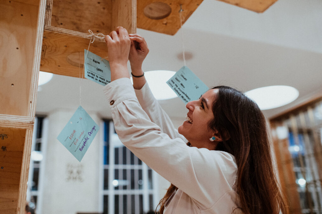 A smiling woman ties a blue card with writing onto a plywood structure indoors, part of a Community Build Project. Other cards hang nearby, reflecting the spirit of youth architecture and collaborative design. The scene is brightly lit, with large windows and circular ceiling lights in the background.