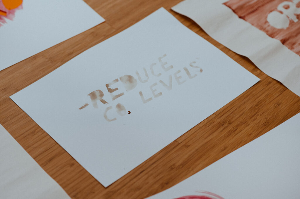 A white paper with cut-out letters reading REDUCE CO2 LEVELS lies on a wooden surface, surrounded by other papers with colourful designs—perhaps concepts for a Community Build Project that encourages young people to design and make structures or sculptures from reused plywood.