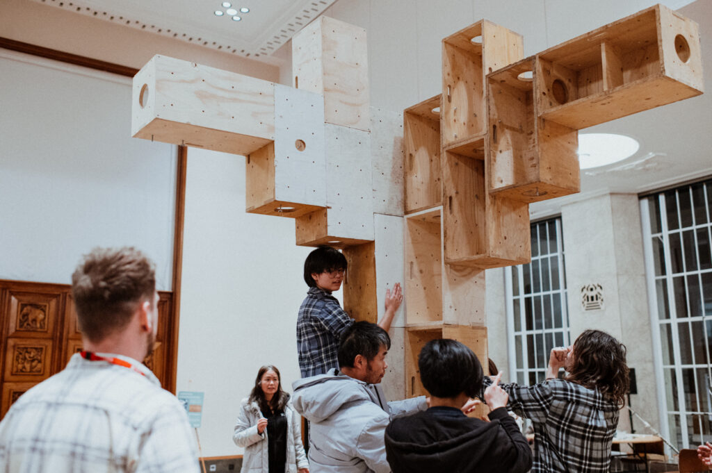 A group of people gather around a tall, geometric wooden structure indoors, some touching or examining the ply sculpture, while others look on or take photos. The scene appears collaborative and creative, capturing the energy of a youth architecture design and community build project focused on making and reusing materials.