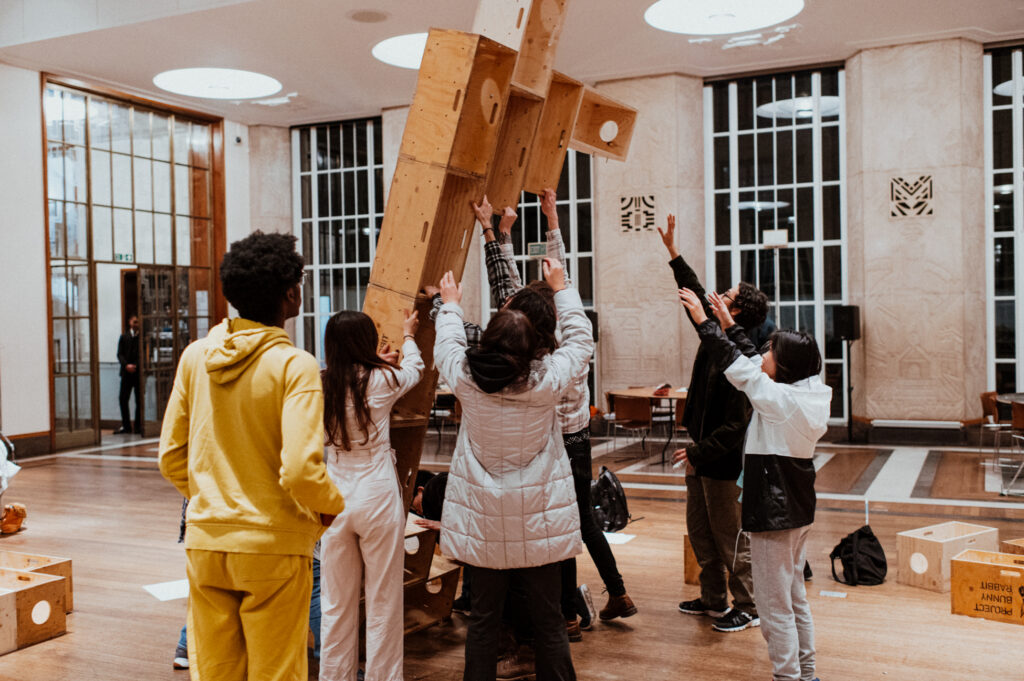 A group of people indoors lift a large plywood structure together as part of a community build project. Some wear jackets and casual clothes. The room has high ceilings, wooden floors, large windows, and geometric wall decorations, creating an inspiring atmosphere for architecture design and sculpture making.