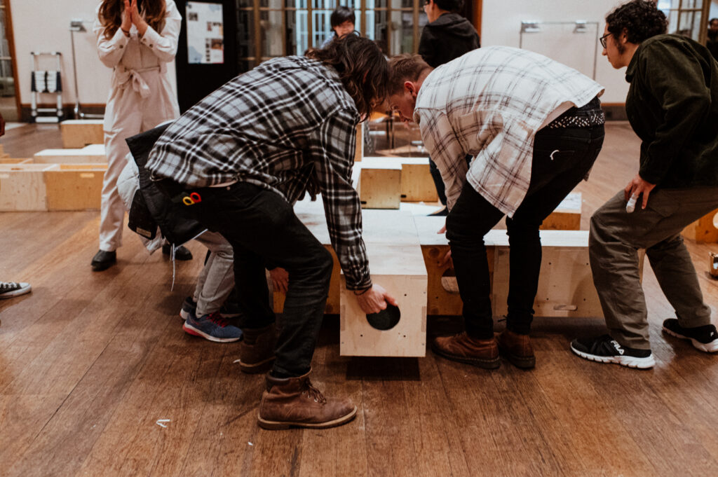 A group of people in casual clothes bend over and interact with wooden blocks on a wooden floor indoors, actively participating in a community build project. Nearby, another person stands taking a photo as the group explores creative design and makes structures using reused plywood.