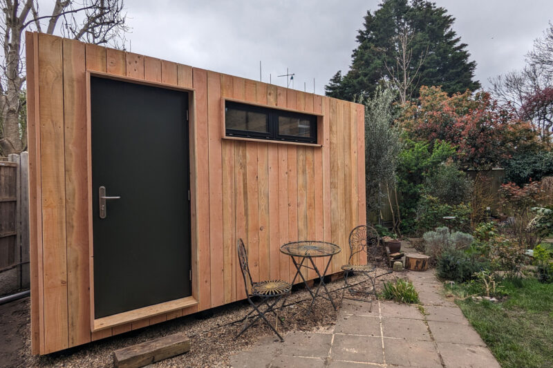 A small birch timber construction garden office with a black door and horizontal window sits on a stone patio. An outdoor metal table and two chairs are nearby, surrounded by plants and trees in this sustainable back garden setting.