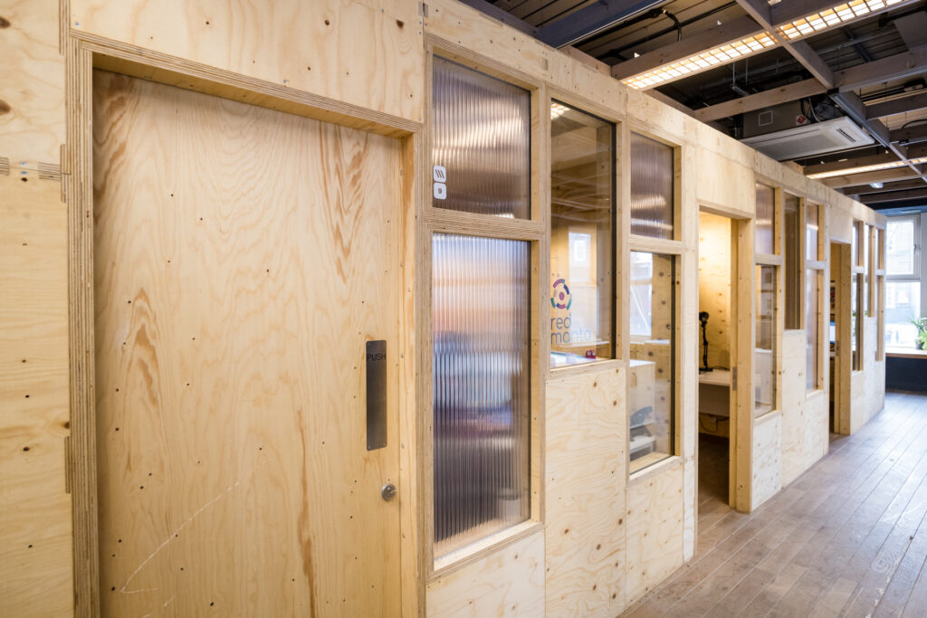 A corridor with timber construction featuring wooden walls and doors, some with ribbed glass windows. Birch accents add warmth to the corridor lined with small offices, one bearing a visible logo. The floor is wooden and the ceiling has exposed fittings.