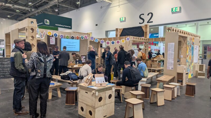People gather inside a spacious, well-lit exhibition hall featuring sustainable wooden furniture and structures made from birch and spruce. Informational posters line the walls, with groups of people seated or chatting near a presentation screen.