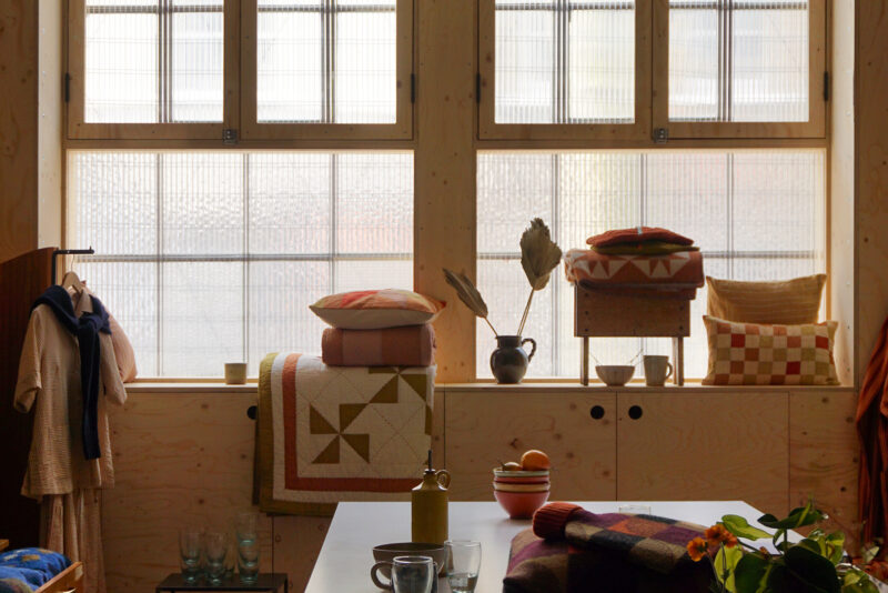 A cosy room with large windows, folded quilts and pillows on a sustainable spruce bench, a coat on a hook, mugs and glasses on a white table, and a vase with dried leaves. Warm, natural light fills the space.