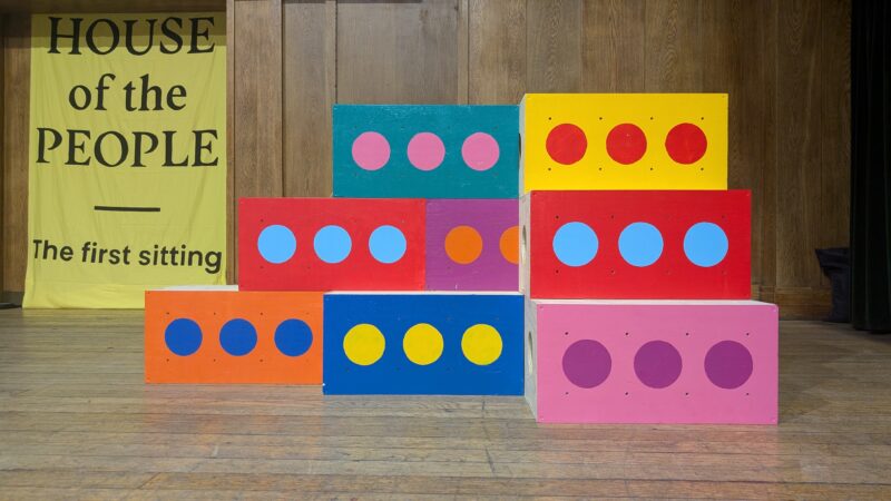 Colourful blocks with circles, made from sustainably sourced materials, are stacked on a wooden floor in front of a yellow banner that reads HOUSE of the PEOPLE - The first sitting, against a wood-panelled wall.