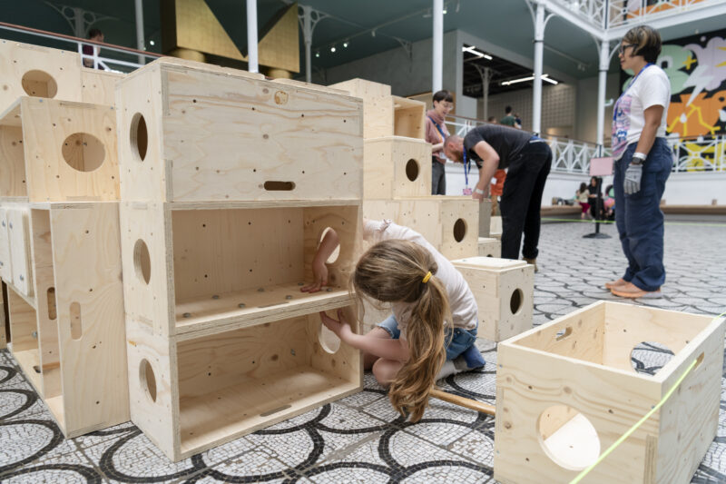 A young girl with long hair builds with large U-Build wooden boxes in an indoor space. Other people work nearby on a tiled floor, surrounded by more wooden structures and colourful wall art.