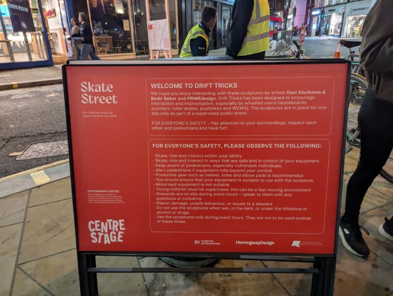 A red U-Build sign titled Skate Street provides safety guidelines and instructions for using drift trick sculptures, with people and a busy street visible in the background at night.