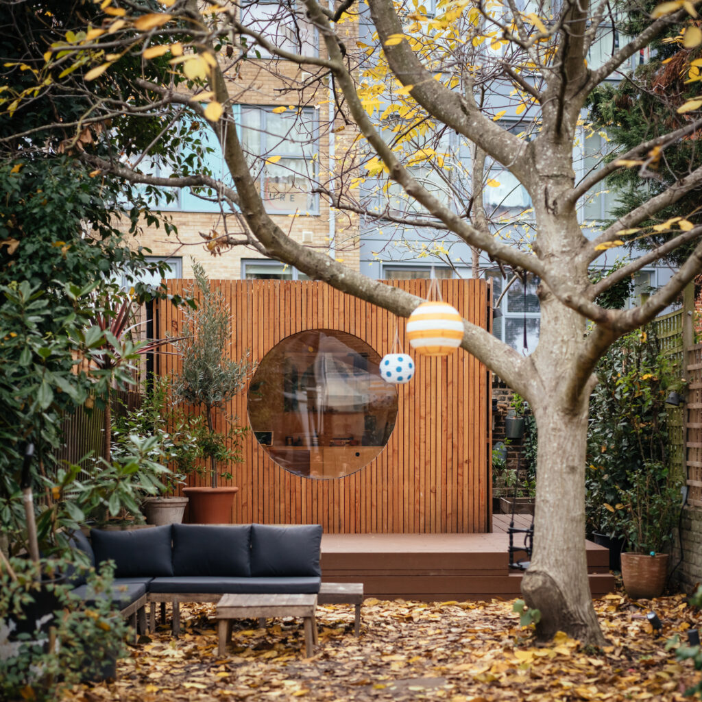 A cosy back garden with a wooden shed featuring a large circular window, surrounded by plants and autumn leaves. Discover why building a Garden Studio could be a fantastic decision for 2026 as you relax on the grey outdoor sofa under hanging decorations.