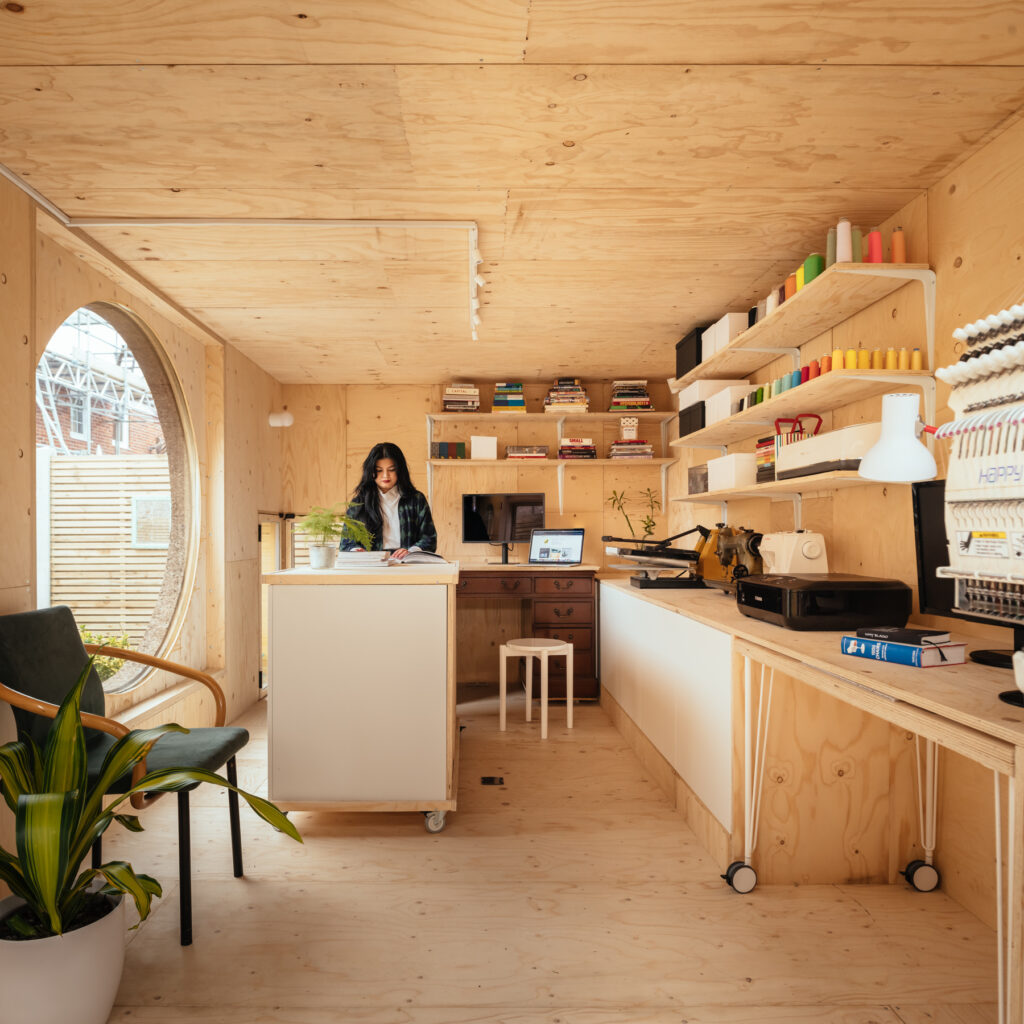 A woman stands at a worktop inside a bright, modern Garden Studio with wooden walls, shelves of colourful spools and books, and a large round window. Plants and office equipment decorate the creative workspace, perfect for building a Garden Studio in 2026.
