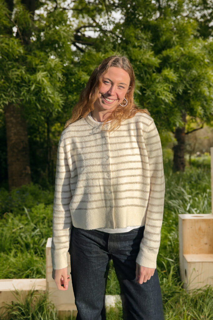 A woman with wavy hair in a cream and beige striped jumper and dark jeans smiles outdoors in a lush green area, reflecting the spirit of sustainable living and inspiring others to build a better future.