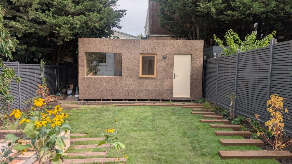 A modern Garden Studio with a cork exterior, window, and door stands at the end of a grassy back garden, bordered by grey fences, flowers, and trees. Paved stepping stones lead across the lawn—an ideal setting for building a Garden Studio in 2026.