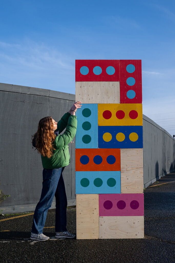 A woman in a green jacket and jeans reaches up to touch a tall, colourful wooden structure made of domino-like pieces, capturing a playful moment on the pavement outside what appears to be a Box Library under the blue sky.