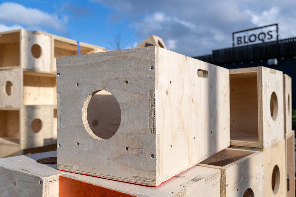 Several unfinished wooden boxes with round cut-outs are stacked outdoors in sunlight, resembling a Box Library. A building with a BLOQS sign is visible in the background under a partly cloudy sky.