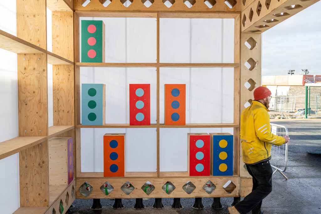 A person in a yellow jacket and red hat stands beside a box library, its wall adorned with colourful rectangular blocks with dots in rows and columns. The outdoor photoshoot takes place on a sunny day, capturing a vibrant moment for 2026.