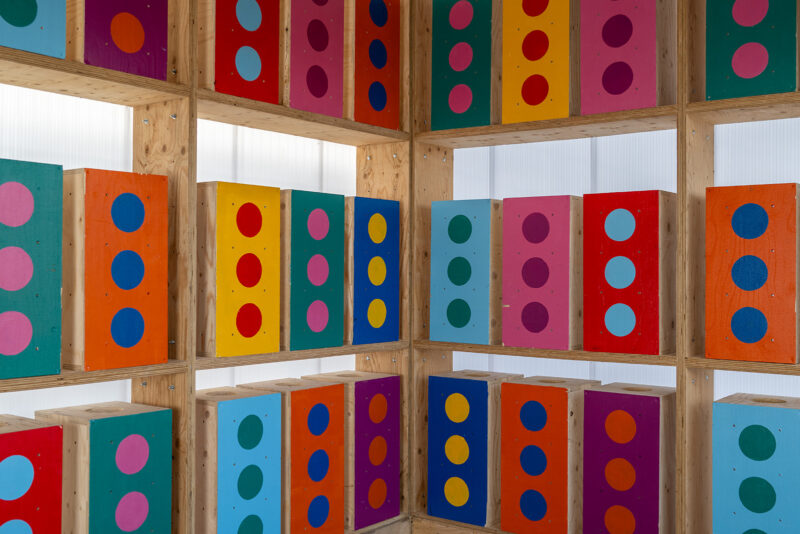 Colourful wooden panels with large painted dots in various colours are arranged on wooden shelves, capturing a vibrant, geometric pattern that covers two walls in this well-lit box library.