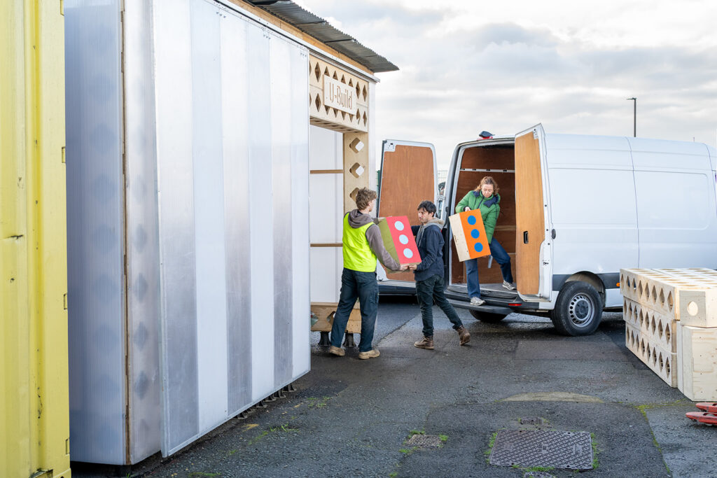 Three people unload colourful wooden panels with circular cut-outs for the Box Library from a white van outside, surrounded by large crate-like structures. One person sits in the van while two carry panels together, capturing a moment of teamwork.