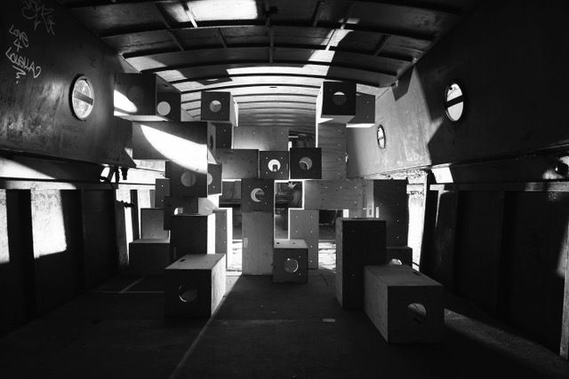Black and white photo of an industrial interior houses a striking installation: stacked rectangular boxes with circular cut-outs. Sunlight streams through openings, casting geometric shadows across the floor in this floating gallery scene.