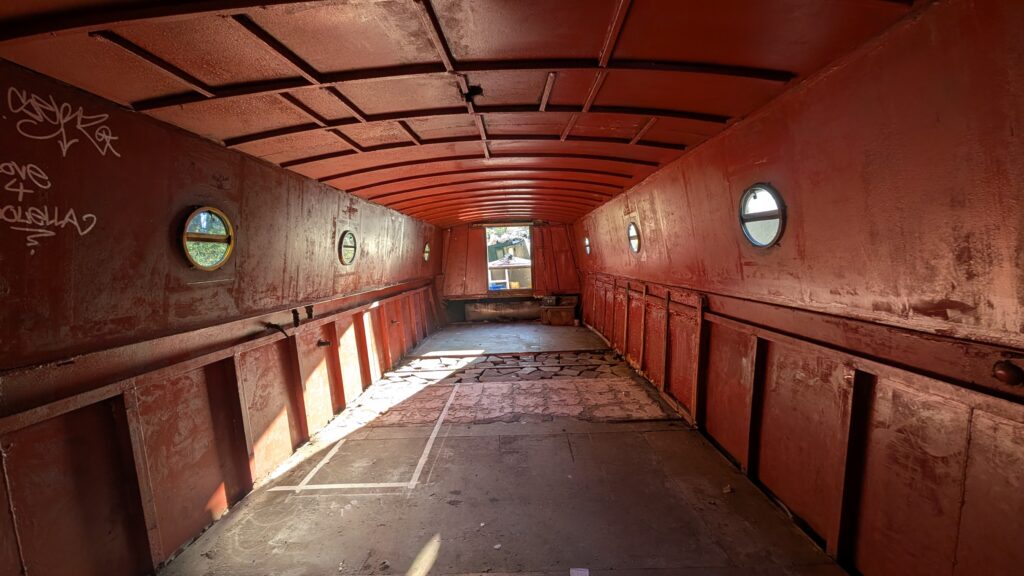 A wide, interior perspective of the empty hull before the installation. The walls and arched ceiling are painted a matte, rustic red, with several small circular porthole windows and white graffiti on the far left. The floor is unfinished concrete with white tape markings, and a doorway at the far end opens to bright daylight and a glimpse of an outdoor setting.
