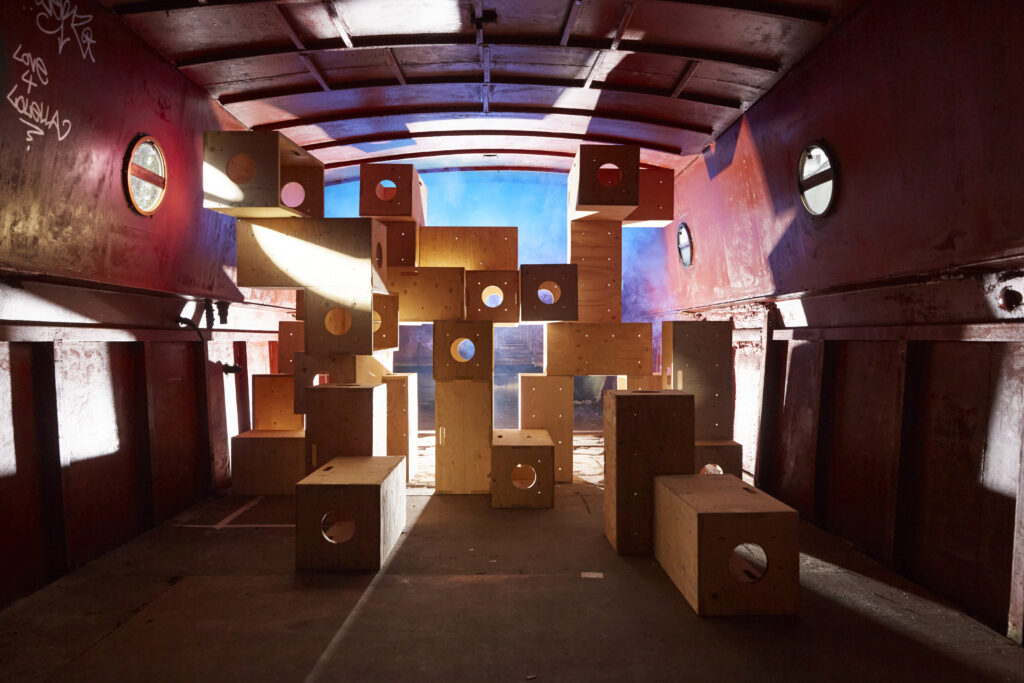 A cluster of modular plywood boxes of various sizes stacked in the center of a red-walled industrial space or cargo container. Bright, warm sunlight streams from the left, casting sharp shadows, while a cool blue light glows from behind the structure. Each wooden box features a circular cutout.