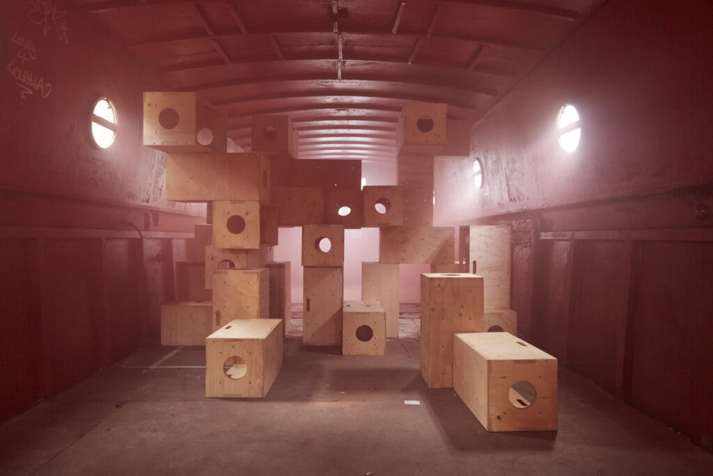 The central stack of plywood modules, now captured in a soft, hazy atmosphere. The lighting is diffused, pink and warm, giving the scene a dreamy, monochromatic red and gold quality. The circular cutouts in the boxes align to create a sense of depth through the center of the stack.