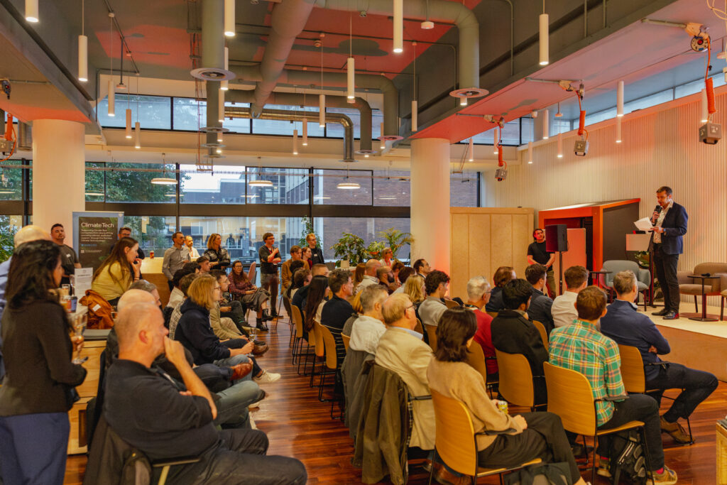 A diverse audience sits in rows facing a stage in Manchester, where a man at the lectern speaks about sustainable business. The modern room has large windows, bright lighting, and people are attentively listening to the Sustainable Ventures presentation.