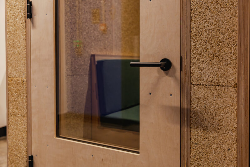 A close-up of a closed wooden door with a glass window and a black handle, revealing a small interior room with a blue cushioned seat inside—an inviting space in Manchester designed by Sustainable Ventures, with textured walls and a natural finish.