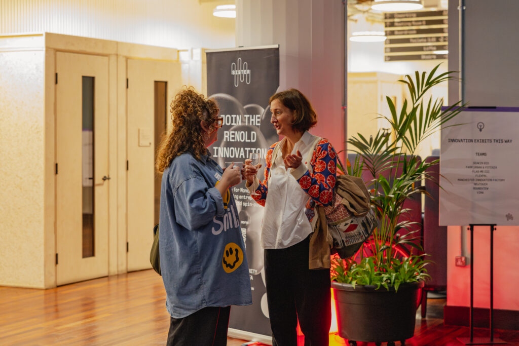 Two women stand and talk at a Manchester indoor event hosted by Sustainable Ventures, with plants and event signs around them. One wears a blue jacket with yellow designs, while the other sports a colourful jacket and a rucksack.