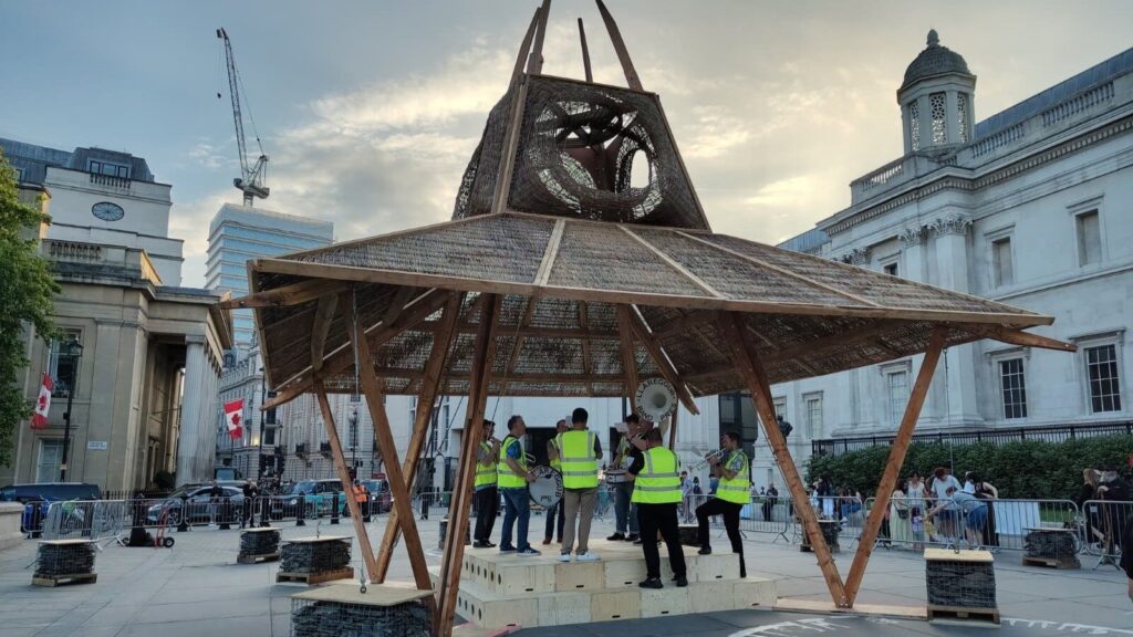 A group of musicians in yellow safety vests perform under a large wooden pavilion in a city square at sunset, surrounded by historic buildings and a crowd celebrating the Triumph of Art, echoing the spirit of the National Gallery’s 2025 festivities.