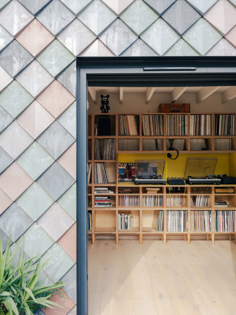 A view through an open door reveals a cosy Record Room in East London, with shelves of vinyl records, a turntable, audio equipment, and pastel diamond-patterned tiles on the exterior wall. A plant sits at the bottom left.