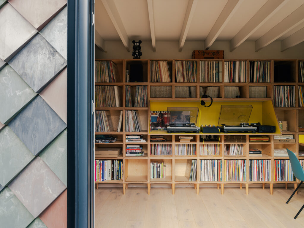 A cosy Record Room in East London with wooden shelves filled with vinyl records, a yellow record player, speakers, and stylish decor. The neatly organised shelves sit beside an exterior wall with grey-green tiles on the left.