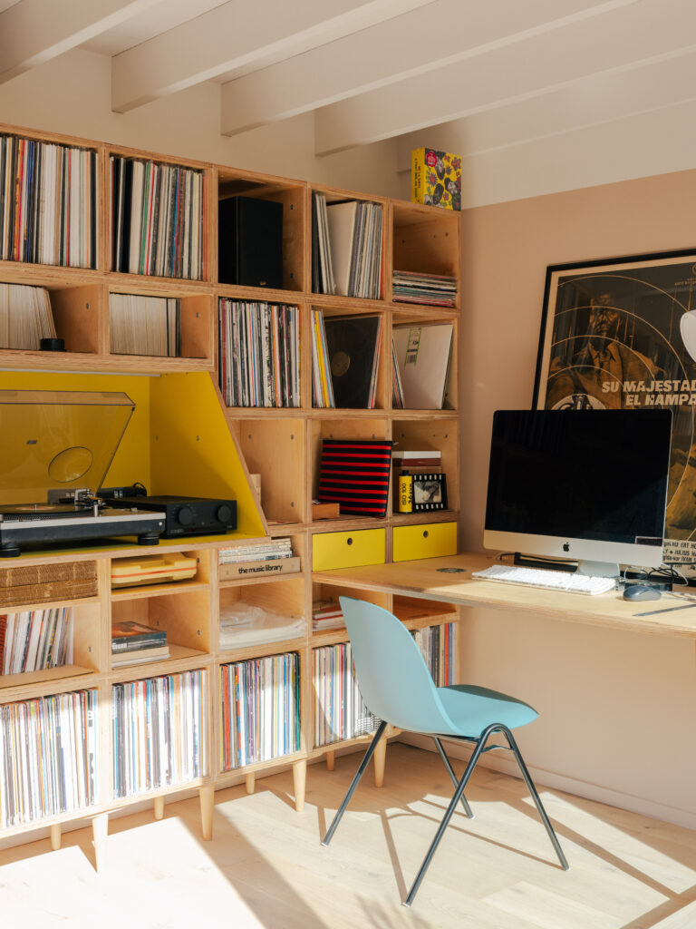 A cosy Record Room home office in East London features a wooden bookcase filled with vinyl records, a turntable, a computer on a built-in desk, a blue chair, and sunlight streaming in through the window.