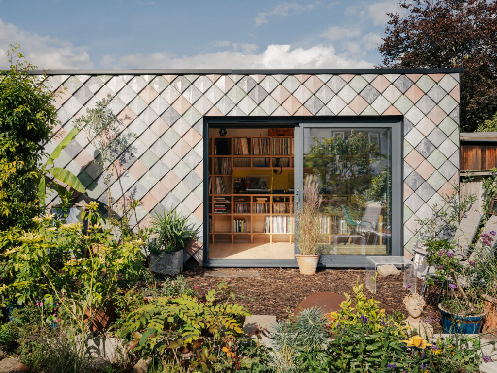 A modern East London garden shed with diamond-patterned exterior tiles and a large sliding glass door, surrounded by lush greenery, reveals a cosy Record Room interior complete with bookshelves and a desk.