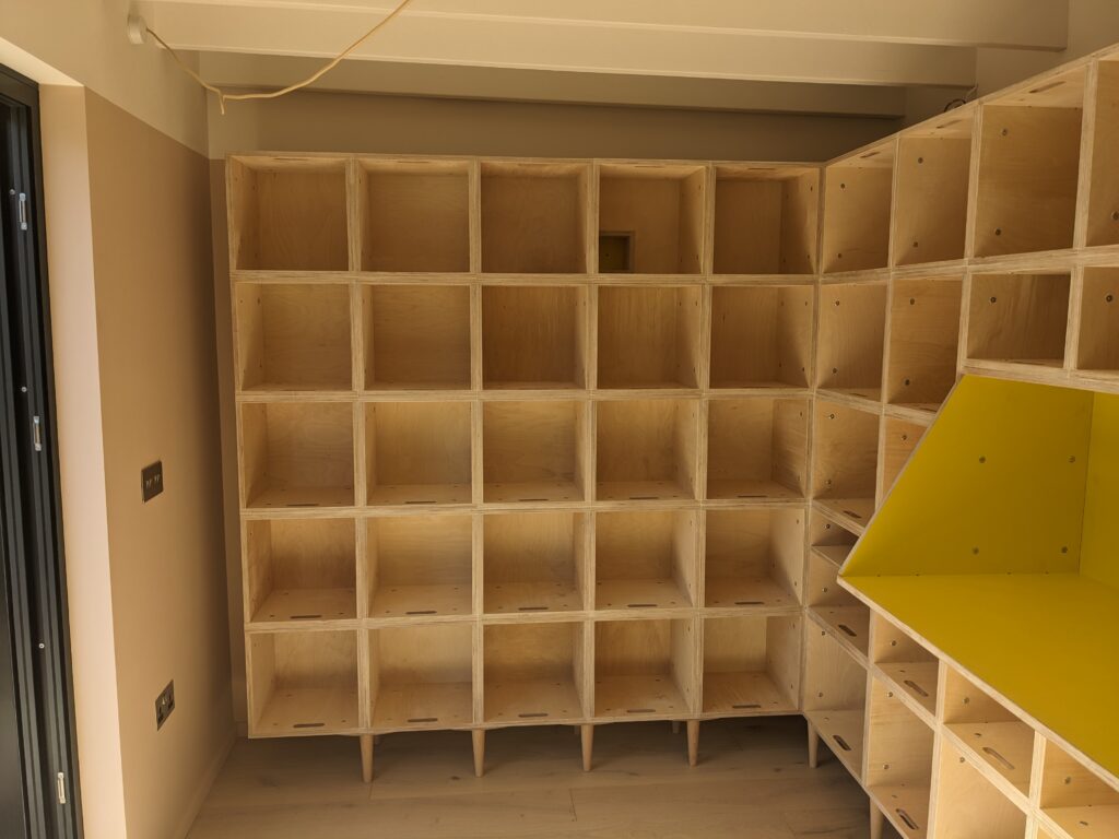 Wooden shelving unit with multiple square cubbyholes fills two walls of this East London Record Room. One section includes a shelf with a bright yellow interior. The room has light walls, a light wooden floor, and exposed ceiling beams.