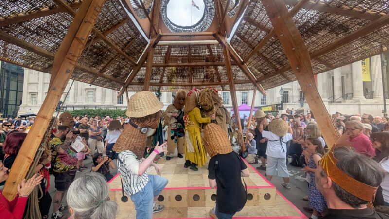 A crowd watches performers wearing large woven straw masks and hats under a wooden pavilion in an outdoor public square, as people take photos and clap—capturing the spirit of the 2025 Triumph of Art at the National Gallery.