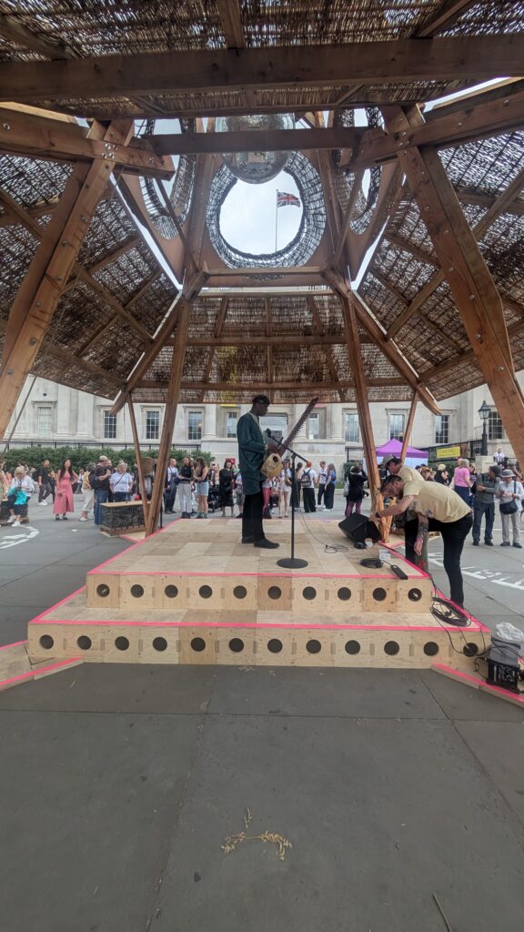 A person stands on a wooden stage under a large, dome-shaped wooden structure at the 2025 Art Exhibition, speaking into a microphone. Another person adjusts equipment nearby as a crowd gathers in the background at the National Gallery.
