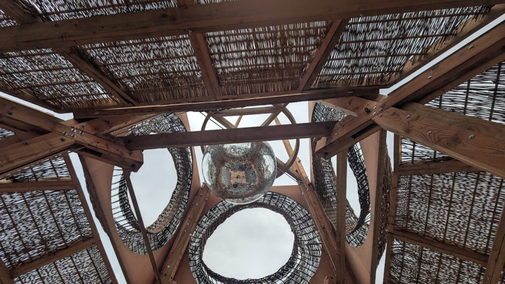 View looking up at a wooden structure with woven panels and circular openings, featuring a large mirrored sphere in the centre—a striking installation at the National Gallery celebrating the Triumph of Art beneath a cloudy sky.
