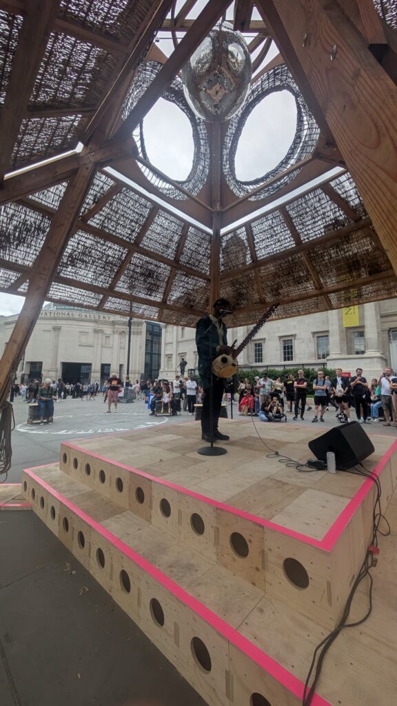 A performer stands on a wooden stage with a microphone, under a woven dome canopy, as part of the Triumph of Art event at the National Gallery. People gather outside, with large buildings in the background beneath a cloudy 2025 sky.