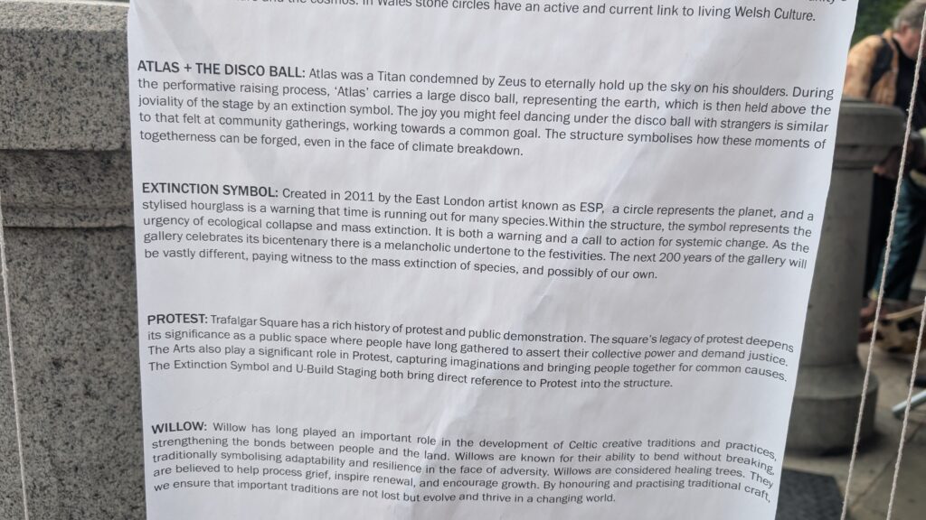A close-up of an informational sign describing four art installations—Atlas + The Disco Ball, Extinction Symbol, Protest, and Willow—featured in the 2025 Triumph of Art event at the National Gallery, attached to a stone surface outdoors.