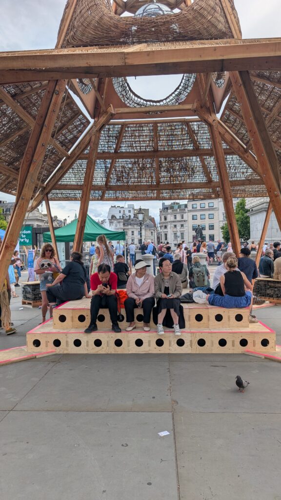 People sit and gather under a large wooden pavilion with a woven roof in a public square. Near the National Gallery, several people enjoy tiered benches or stroll by, embracing the spirit of "Do Your Own Thing." Green marquees and buildings frame the scene.